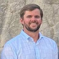Portrait of Garrett Fell, President of B.G. Hoadley Quarries Inc., standing in front of a light stone background, wearing a light blue button-up shirt and smiling.