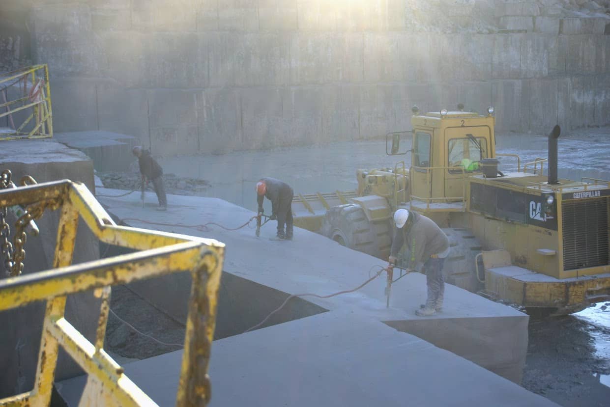 Three quarry workers in safety gear cutting large limestone blocks with pneumatic tools inside B.G. Hoadley’s quarry pit, with a Caterpillar loader nearby in soft morning light.