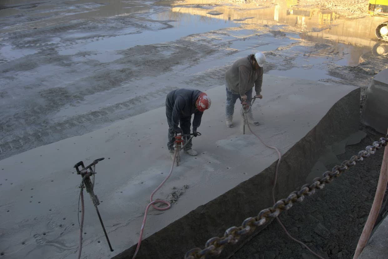 Two quarry workers using pneumatic drills to perforate the surface of a large limestone block inside a muddy quarry floor at B.G. Hoadley Quarries.