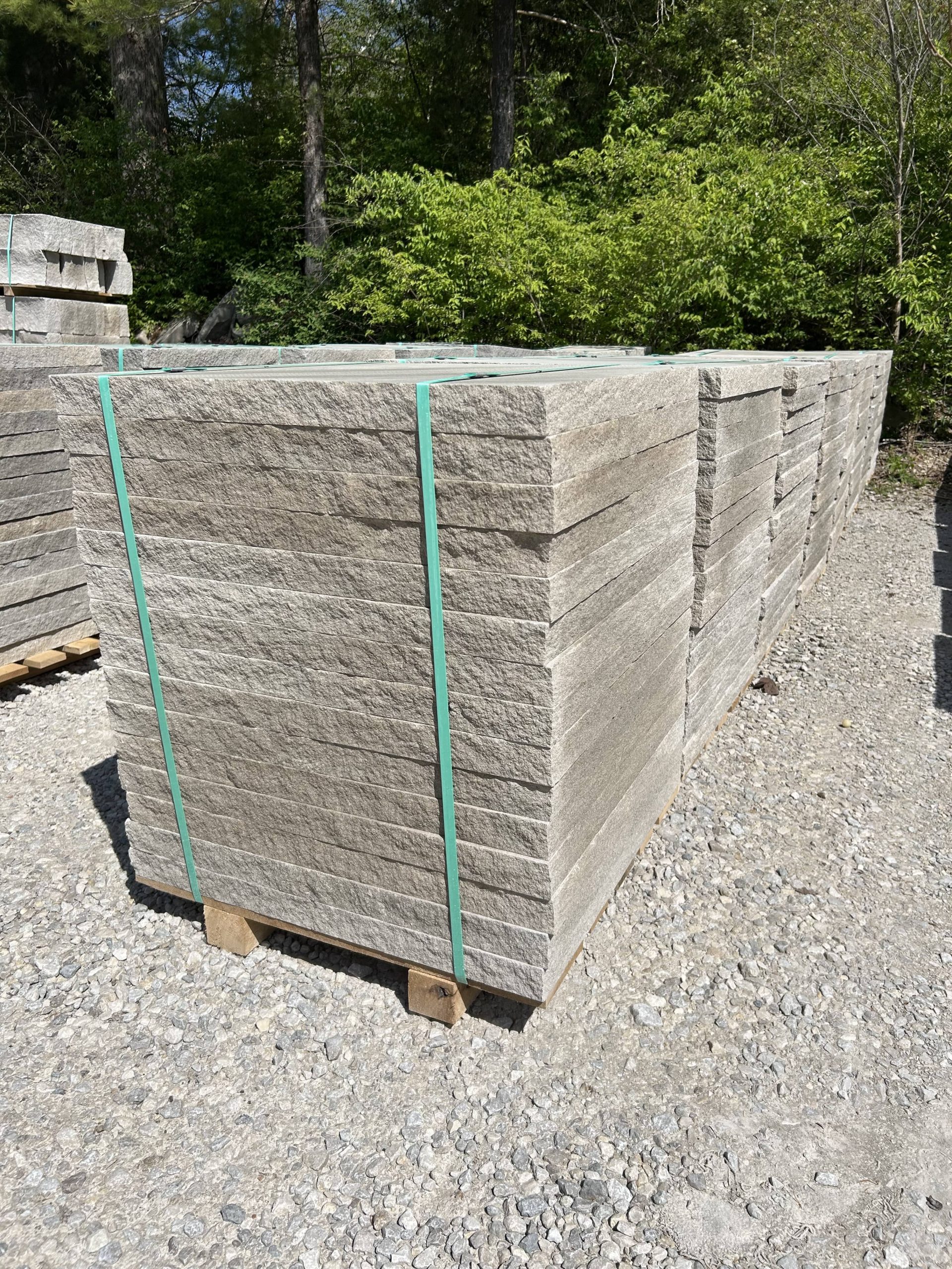 Close-up of a pallet of natural stone pavers stacked in even layers and strapped with green bands, placed on gravel with greenery in the background.
