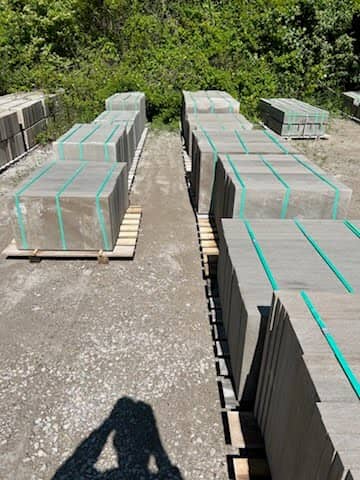 Top-down view of concrete pavers strapped and stacked on wooden pallets in an outdoor gravel yard with green trees in the background.