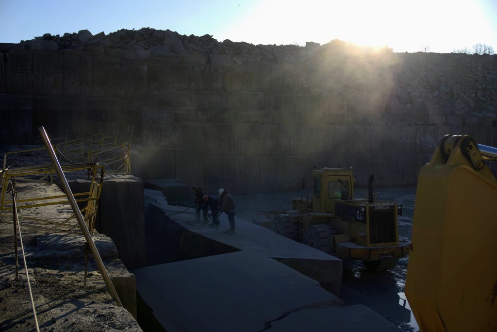 Wide-angle view of a terraced limestone quarry at sunrise with silhouetted workers using tools and heavy Caterpillar machinery in the foreground