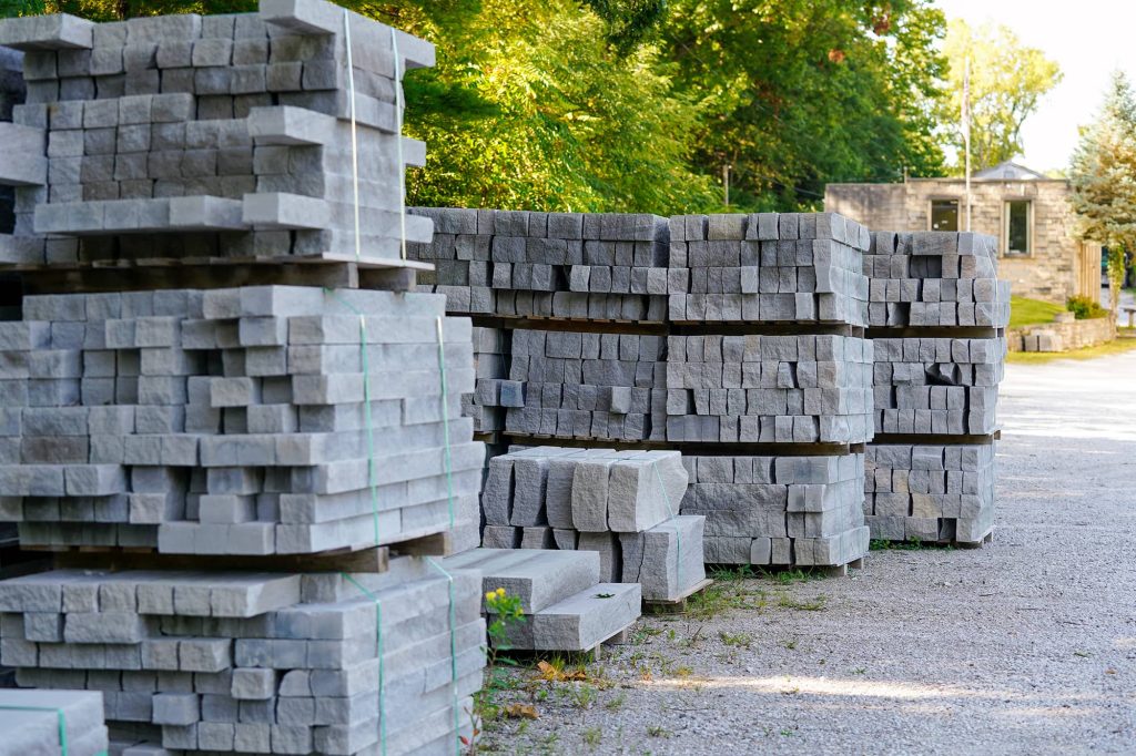 Stacks of split-face ashlar limestone blocks organized on pallets in a gravel industrial yard with a stone building in the background.