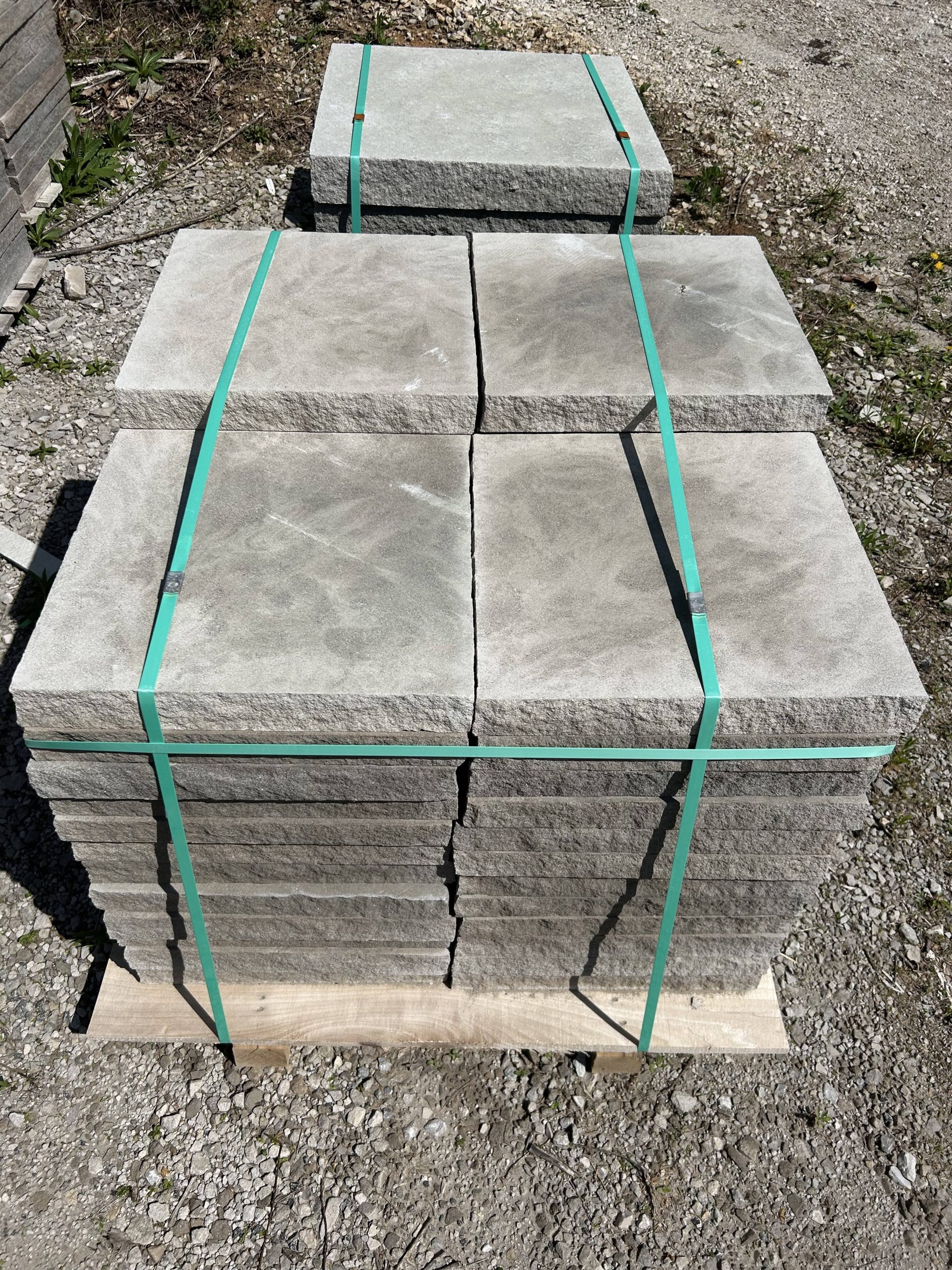 A bundle of natural stone pavers neatly stacked on a wooden pallet, secured with green plastic straps, placed on gravel ground.