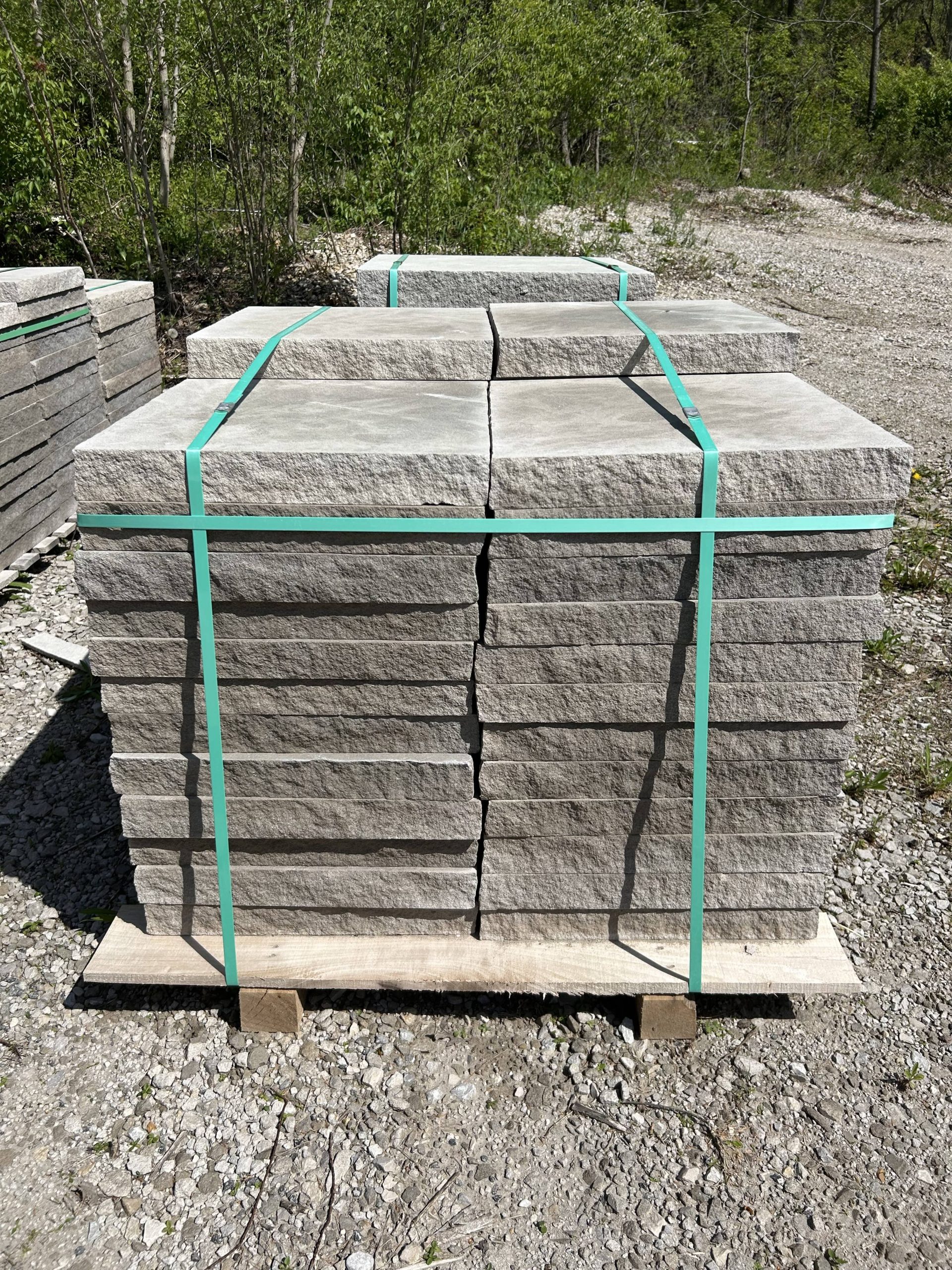 Front-facing view of a tightly stacked pallet of natural stone pavers, strapped with green bands and set on gravel with a forested background.