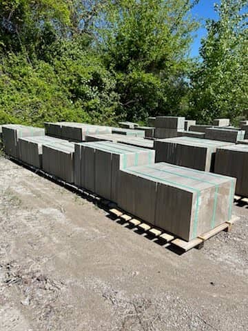 Concrete pavers stacked and strapped on wooden pallets, arranged in rows in an outdoor dirt yard with green trees in the background.
