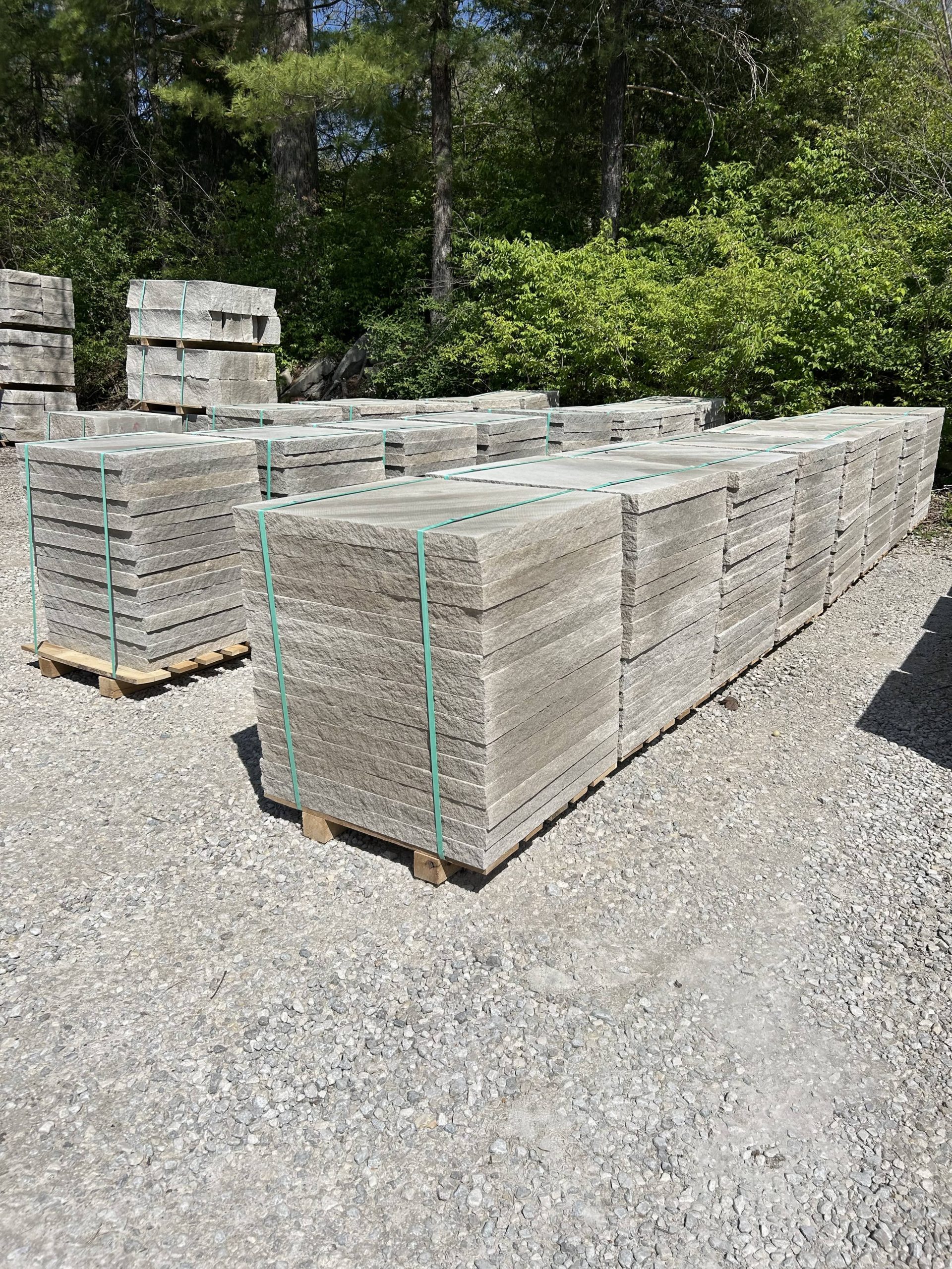 Several large pallets of natural stone pavers stacked and secured with green straps, stored on a gravel lot with a forested backdrop.