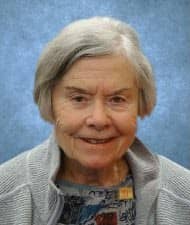 Portrait of Patsy Fell-Barker, an elderly woman with short gray hair, smiling warmly while wearing a gray sweater against a blue textured background.