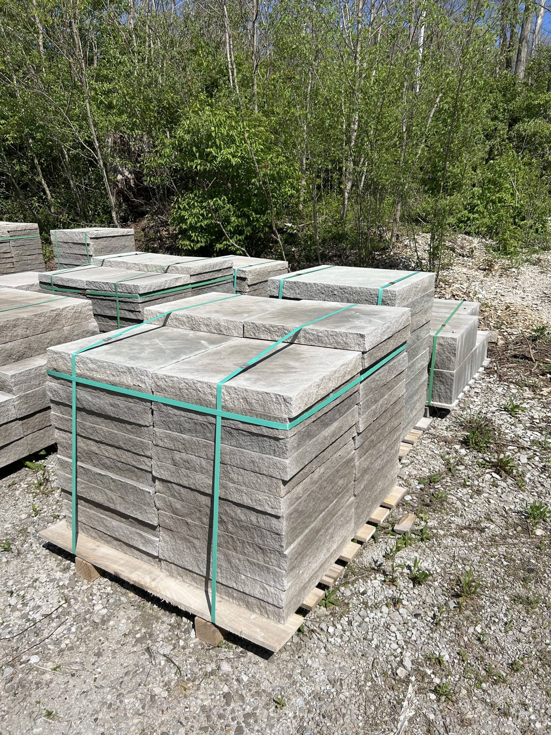 Pallet of square-cut natural stone pavers stacked and secured with green straps on a gravel lot near a wooded area.
