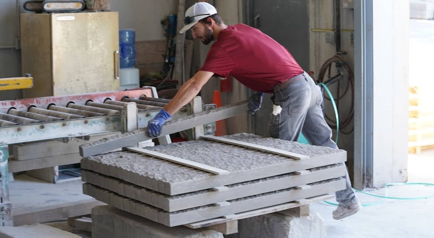 Worker in red shirt and cap loading chiseled stone slabs onto a conveyor in a stone fabrication workshop.