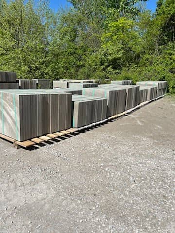 Multiple stacks of large concrete pavers secured with straps, arranged on wooden pallets in an outdoor gravel yard surrounded by greenery.