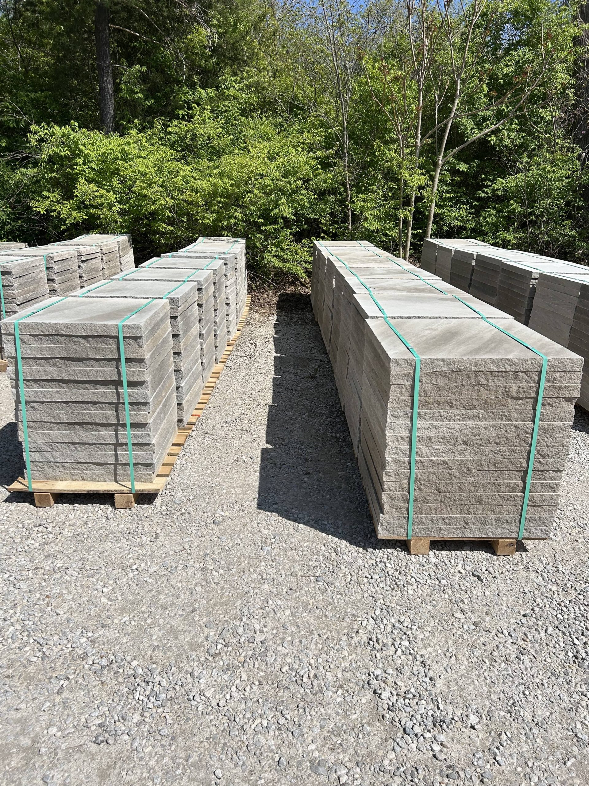 Two rows of stacked stone pavers strapped with green bands on wooden pallets, placed on a gravel surface beside dense green trees.