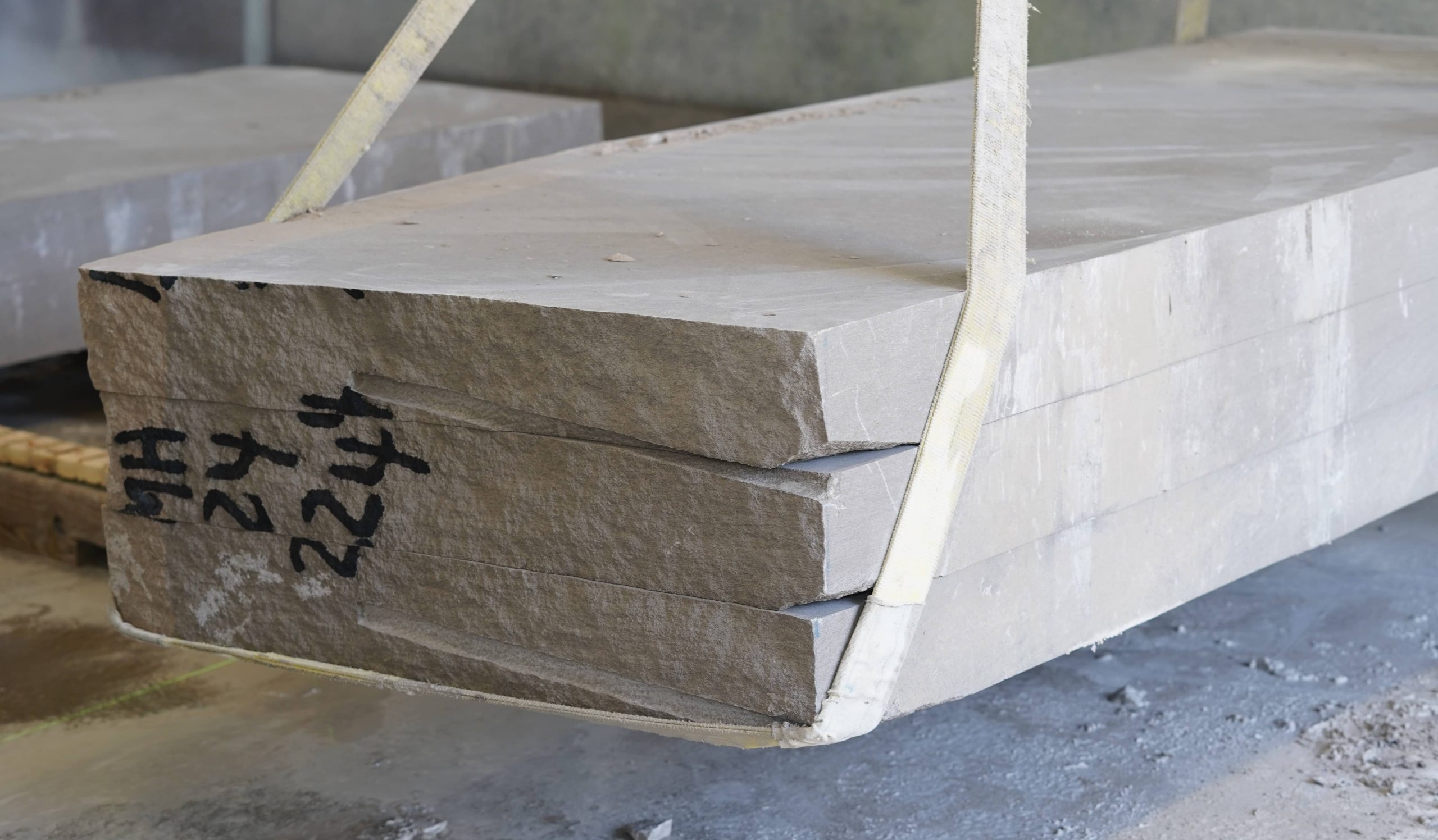 A stack of 2-inch thick variegated limestone slabs suspended by lifting straps inside a stone fabrication facility.